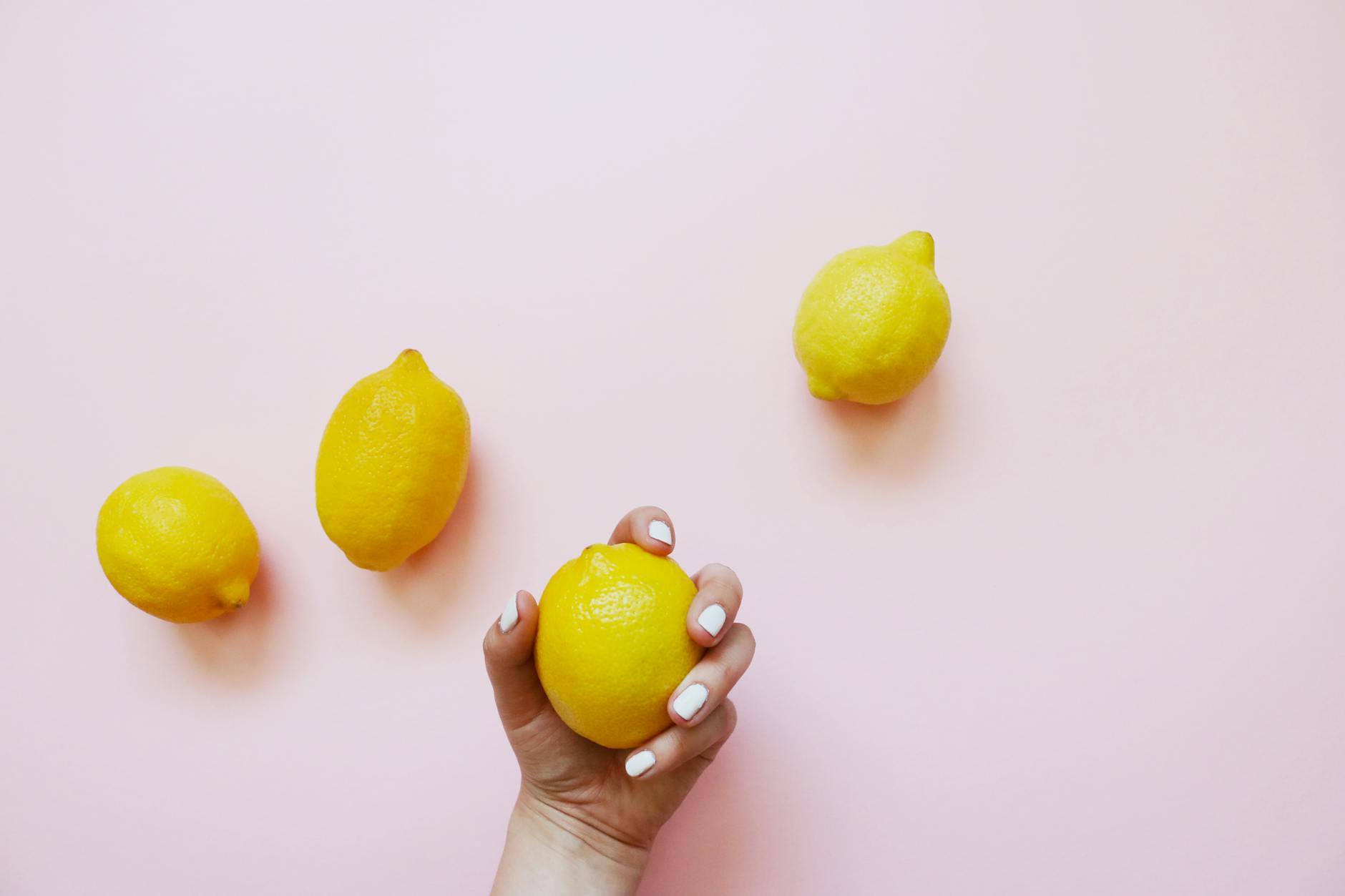 A hand holding a lemon on a soft pink background with lemons scattered nearby